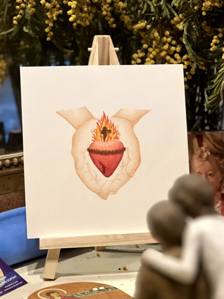 Un Sacré-Coeur surmonté d'un feu brûlant et d'une croix dans ce feu, entouré de la couronne d'épines, avec la plaie au côté droit, tenu dans les mains de Jésus, pour symboliser l'Amour qu'Il nous offre.
L'aquarelle est posée sur un trépied, dans le coin prière familial.